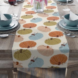 May include: Cream-colored table runner featuring a repeating pattern of stylized pumpkins in shades of orange, green, pink, and blue. Each pumpkin has the text "fuck trump" written on it. The runner is placed on a wooden table.
