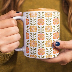 May include: White ceramic mug with a floral pattern in orange, yellow, and pink. The design includes the text "fuck trump" repeated vertically. The mug is held by a person with dark blue nail polish.