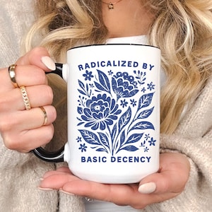 May include: White ceramic mug with a black handle and rim. The mug features a blue floral design and the text "RADICALIZED BY BASIC DECENCY". The mug is held in a person's hands.