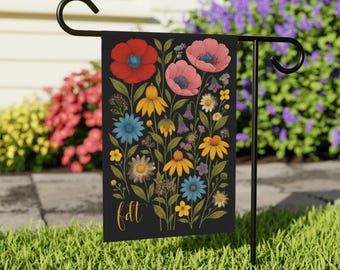 May include: A black garden flag featuring a floral design with red poppies, pink flowers, yellow daisies, and blue flowers. The word "fall" is written in gold script at the bottom. The flag is displayed on a black metal stand in a grassy yard.
