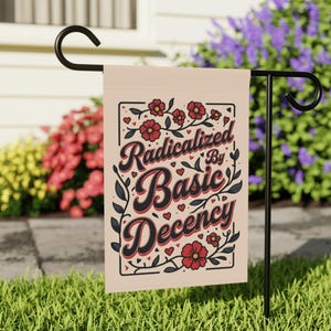May include: A beige garden flag with the text "Radicalized By Basic Decency" in a decorative font, framed by red flowers, black leaves, and small hearts. The flag is hanging on a black metal stand in a garden setting.