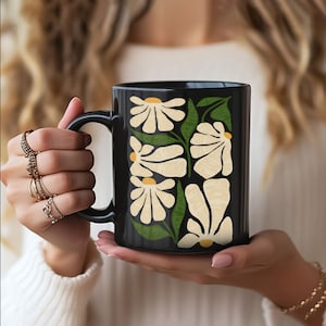 May include: Black ceramic mug featuring a floral design with cream-colored flowers, yellow centers, and green leaves. The mug has a black handle and is held by a person wearing rings and a bracelet. The mug is a standard size.