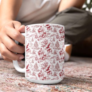 May include: White ceramic mug featuring a red and white toile pattern with Santa Claus figures, trees, and other holiday elements. The mug has a large handle and a wide base, perfect for hot beverages.