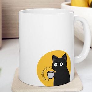 May include: White ceramic mug featuring a black cat illustration on a yellow circle. The cat holds a cup of coffee, with the text "IS HE DEAD YET?" around the circle. The mug sits on a beige coaster.