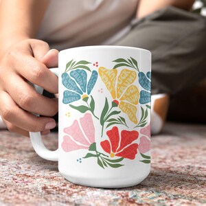 May include: White ceramic mug featuring a floral design with the words "FUCK TRUMP" in a repeating pattern within the flower shapes. The flowers are in shades of blue, yellow, pink, and red, with green leaves.