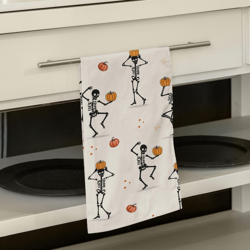 May include: White Halloween kitchen towel with a repeating pattern of black skeleton figures holding orange pumpkins. The text "FUCK" is printed on the skeletons. Small orange pumpkins and stars are scattered across the towel.