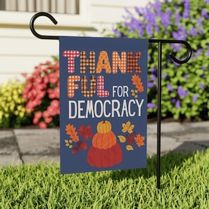 May include: A rectangular navy blue garden flag hanging on a black metal stand. The text THANKFUL FOR DEMOCRACY is printed in white and plaid patterns. Below the text are three stacked pumpkins in yellow, orange, and red with autumn leaves.