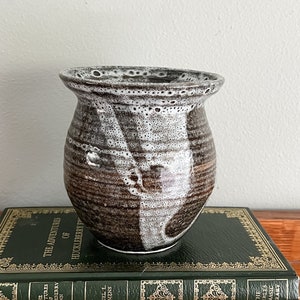 May include: A small, brown and white ceramic vase with a speckled glaze. The vase is sitting on top of two green books.