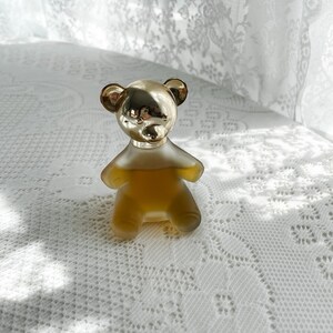 May include: A gold bear-shaped bottle of perfume sits on a white lace tablecloth. The bottle is filled with a clear, amber-colored liquid.