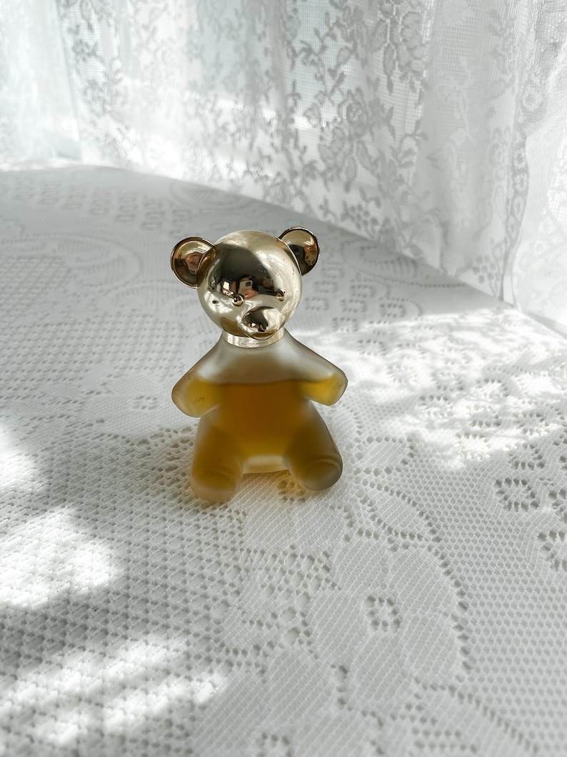 May include: A gold bear-shaped bottle of perfume sits on a white lace tablecloth. The bottle is filled with a clear, amber-colored liquid.