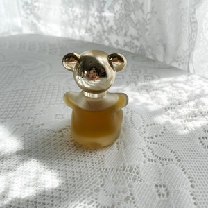 May include: A gold bear-shaped bottle of perfume with a clear glass body and a gold cap. The bottle is sitting on a white lace tablecloth.