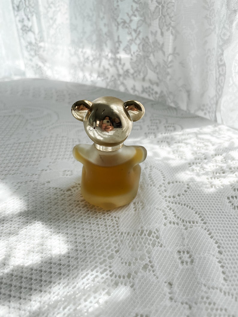 May include: A gold bear-shaped bottle of perfume with a clear glass body and a gold cap. The bottle is sitting on a white lace tablecloth.