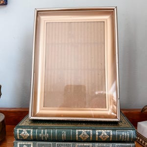 May include: A gold-toned rectangular picture frame with a light brown mat, resting on two vintage green books with gold lettering. The books are titled "Oliver Twist" and "The Adventures of Huckleberry Finn."