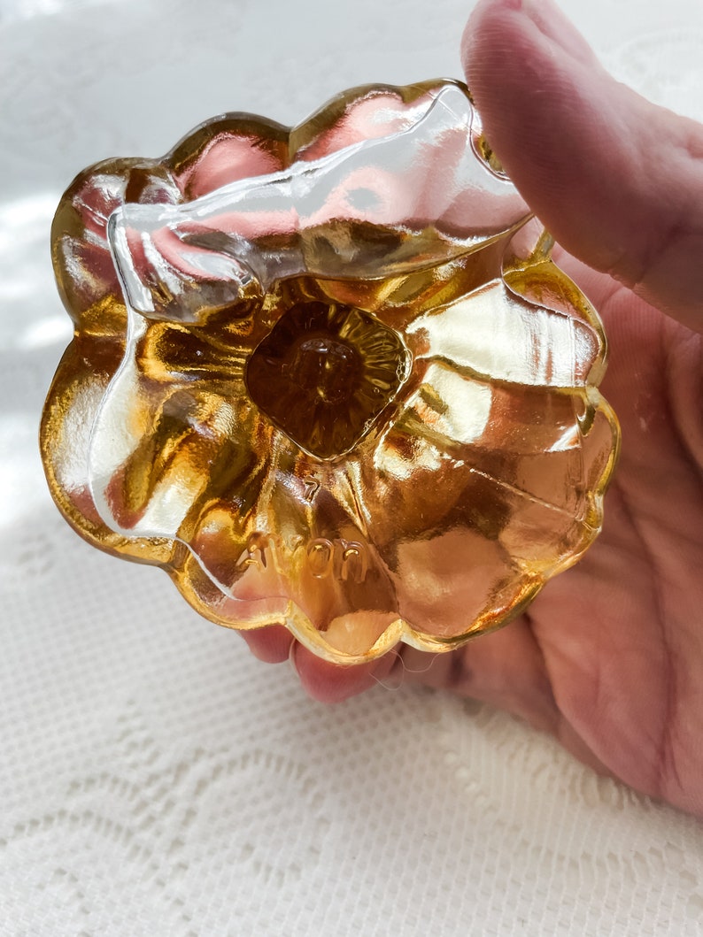 Pode incluir: Um tamp&atilde;o de garrafa de vidro dourado em forma de flor com a palavra "AVON" gravada na parte inferior.