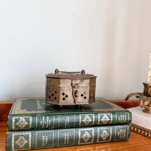 May include: A brass box with a hinged lid and decorative cutouts sits on top of two green hardcover books. The books are titled "The Adventures of Sherlock Holmes" and "Oliver Twist".