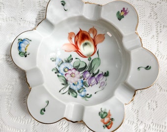 May include: A white porcelain ashtray with a gold rim and a floral design. The flowers are painted in shades of pink, orange, blue, and purple.