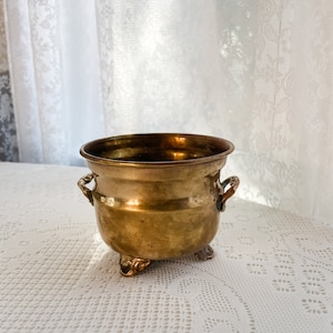 May include: A small, gold-colored brass planter with three feet and two handles. The planter is sitting on a white lace tablecloth.