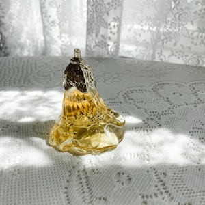 Pode incluir: Um frasco de perfume de vidro transparente em forma de p&ecirc;ra com uma tampa prateada. O frasco est&aacute; cheio de um l&iacute;quido amarelo e est&aacute; sobre uma toalha de renda branca.