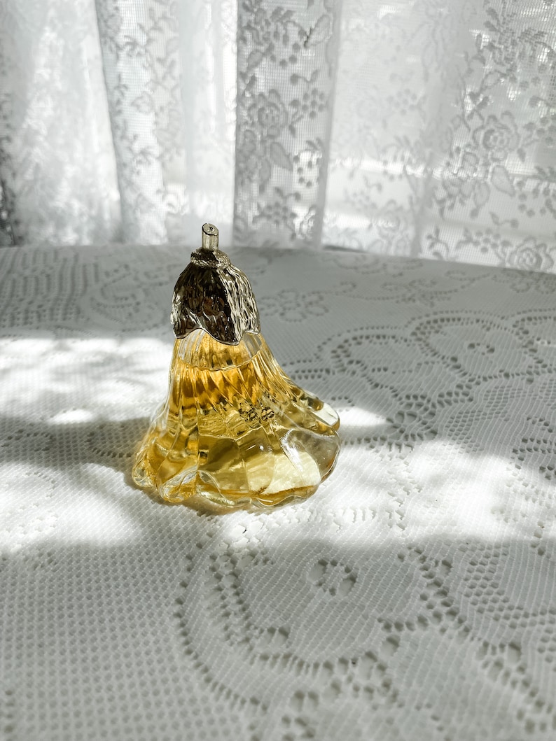 Pode incluir: Um frasco de perfume de vidro transparente em forma de p&ecirc;ra com uma tampa prateada. O frasco est&aacute; cheio de um l&iacute;quido amarelo e est&aacute; sobre uma toalha de renda branca.