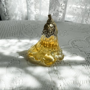 Pode incluir: Um frasco de perfume de vidro transparente em forma de flor com uma tampa dourada. O frasco est&aacute; cheio de um l&iacute;quido amarelo e est&aacute; sobre uma toalha de renda branca.