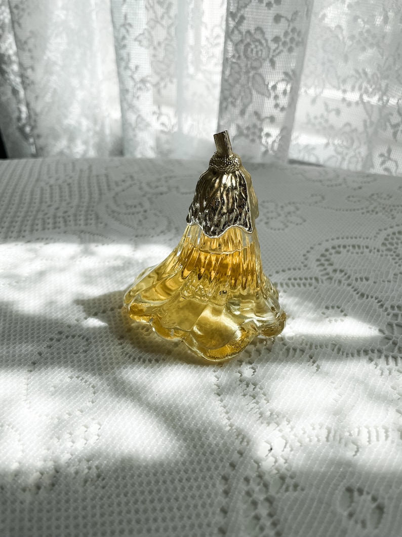 Pode incluir: Um frasco de perfume de vidro transparente em forma de flor com uma tampa dourada. O frasco est&aacute; cheio de um l&iacute;quido amarelo e est&aacute; sobre uma toalha de renda branca.