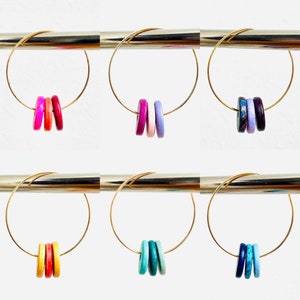 Könnte beinhalten: Goldene Creolen-Ohrringe mit jeweils drei farbigen Holzperlen. Die Perlen sind rund und glatt, in den Farbtönen Rot, Rosa, Lila, Blau und Türkis. Die Creolen sind dünn und filigran, was ein schlichtes, aber auffälliges Design ergibt.