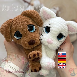 May include: A brown crocheted dog and a white crocheted cat with blue eyes. The dog has brown paws and the cat has pink paws. The text "DADUŠKA BALUCHOVÁ" is visible at the top of the image.