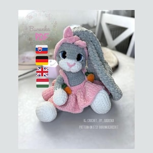 May include: A crocheted gray bunny wearing a pink dress and a pink bow. The bunny is holding two orange carrots. The image includes the text "Barunka PDF" and "IG: _CROCHET_BY_ _DAOUSKA PATTERN ON ETSY BARUNKACROCHET".