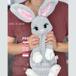 Könnte beinhalten: Ein grauer Häkelhase mit rosa Ohren und weißen Füßen. Der Hase hat große blaue Augen und eine rosa Nase. Der Hase wird von einer Person in einem roten Shirt gehalten. Der Text "PDF" ist im Bild sichtbar. Der Text "IG: CROCHET_BY_DADUSKA" ist im Bild sichtbar. Der Text "PATTERN ON ETSY BARUNKACROCHET" ist im Bild sichtbar.