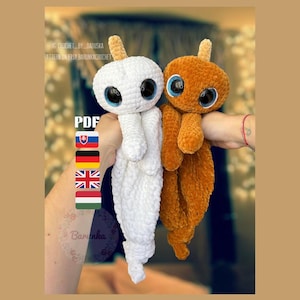 May include: Two crocheted amigurumi toys, one white and one brown, with large googly eyes and a horn on top of their heads. The toys are being held by a person's hands. The text "PDF" is visible in the image.