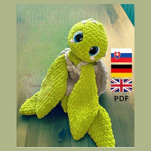 Op de afbeelding: Een handgemaakte, groene gehaakte schildpad met grote, blauwe ogen en een lichtgrijze schild. De schildpad heeft een getextureerd uiterlijk. De afbeelding bevat de tekst "DADUSKA BALUCHOVA" en vlaggen voor Slowakije, Duitsland en het VK.