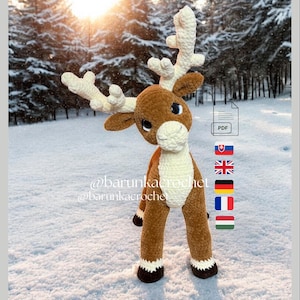 May include: A handmade brown and cream crochet reindeer with large antlers, standing in the snow. The reindeer has a white belly and cream-colored antlers. The background shows a snowy forest. The text "@barunkacrochet" is visible.