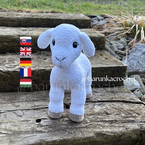 May include: A handmade white crochet lamb with a fluffy texture. The lamb has small black eyes, a stitched nose, and tan hooves. The lamb is standing on a wooden surface. The image includes the text "@barunkacrochet".