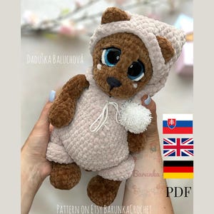 Peut inclure: Ours en peluche crocheté portant une tenue à capuche rose clair et marron. L'ours a de grands yeux bleus et un pompon blanc sur sa patte. Le texte "PATTERN ON ETSY BARUNKA CROCHET" est visible en bas de l'image.