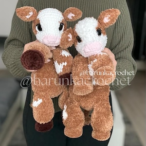 May include: Two handmade crochet cow plushies. The plushies are brown and white with large, dark eyes and pink noses. The ears and hooves are a darker brown. The text "@barunkacrochet" is visible on the image.