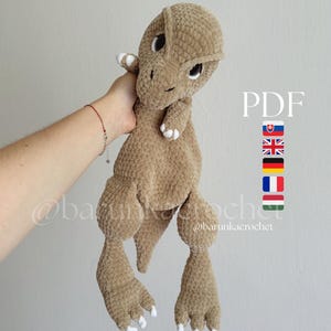 Puede incluir: Un dinosaurio de peluche color canela hecho de hilo suave y texturizado con detalles blancos en las garras. El juguete presenta ojos grandes y oscuros y una cola larga. El texto PDF es visible a la derecha junto a iconos de banderas nacionales.