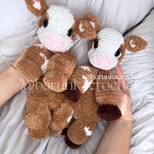 May include: Two handmade crochet cow plushies. The plushies are brown with white patches, pink noses, and black eyes. The ears and hooves are a darker brown. The text "@barunkacrochet" is visible on the image.