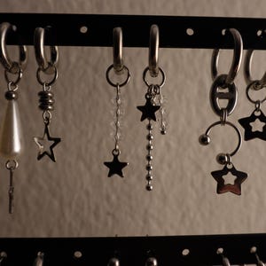 May include: A display of various silver earrings, including hoop earrings and dangle earrings. Some earrings feature star charms, beaded accents, and pearl-like drops. The earrings are displayed on a black rack against a neutral background.