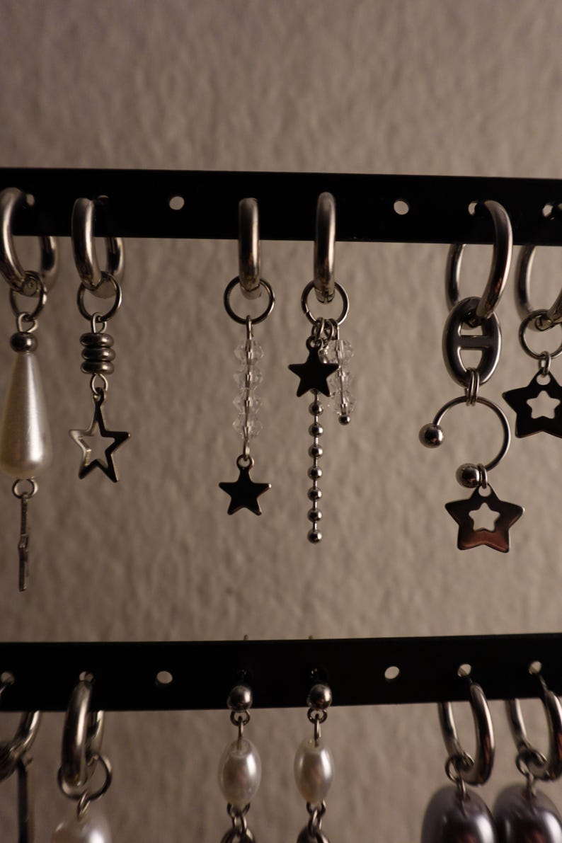 May include: A display of various silver earrings, including hoop earrings and dangle earrings. Some earrings feature star charms, beaded accents, and pearl-like drops. The earrings are displayed on a black rack against a neutral background.