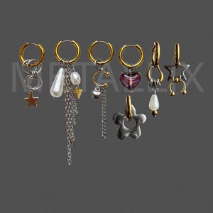 May include: A collection of six gold-toned hoop earrings featuring various dangling charms including a star, a white pearl, a silver chain, a small heart, a purple glass heart, a flower-shaped pendant, and a horseshoe-shaped metal piece.