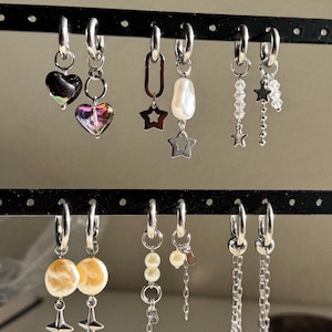 May include: A collection of silver hoop earrings with various charms, including hearts, stars, pearls, and chains. Some earrings feature iridescent heart-shaped pendants, while others have dangling star accents or beaded strands. The earrings are displayed on a black earring holder.