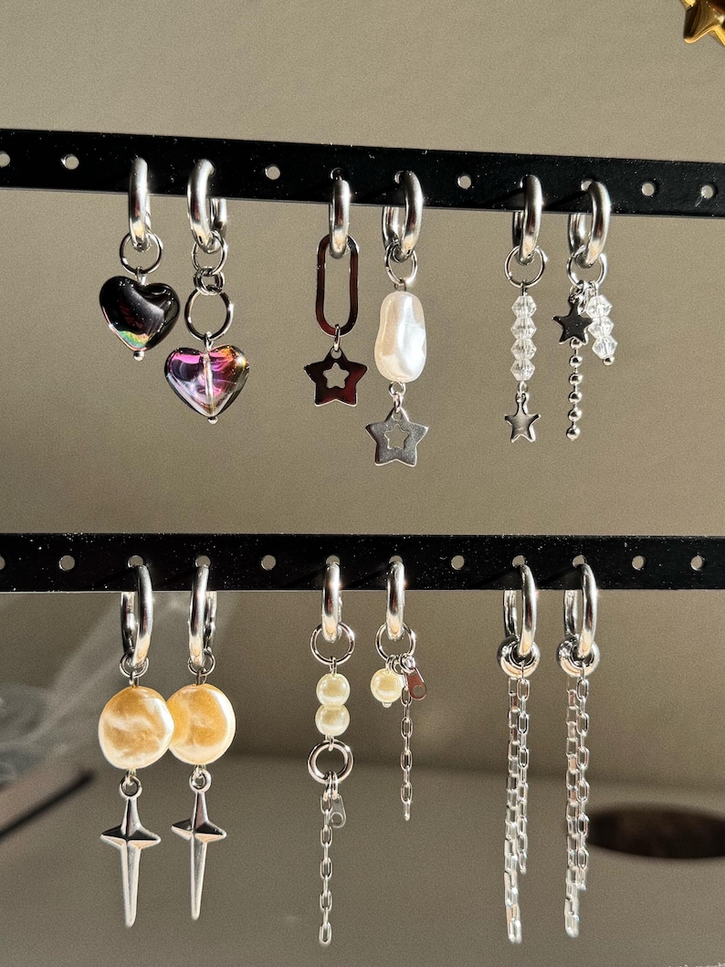 May include: A collection of silver hoop earrings with various charms, including hearts, stars, pearls, and chains. Some earrings feature iridescent heart-shaped pendants, while others have dangling star accents or beaded strands. The earrings are displayed on a black earring holder.