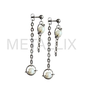 May include: A pair of silver-toned chain earrings. Each earring features a small, round pearl suspended within a circular frame at the bottom, connected by a chain to a smaller pearl and a stud. The word "METALIX" is partially visible in the background.