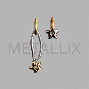 May include: A pair of asymmetrical earrings. One earring features a gold hoop with a silver oval and a gold star charm. The other earring has a gold hoop with a gold star charm. The word "METALLIX" is in the background.