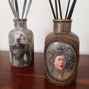 Op de afbeelding: Twee decoratieve glazen flessen met rietverspreiders. Eén fles toont een eend-mens hybride in een vintage jurk, de andere een portret van Medusa. Beide flessen hebben een rustieke, verouderde uitstraling, met donkere rietjes.