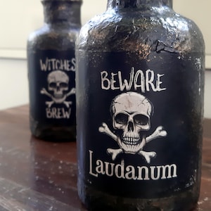 Könnte beinhalten: Zwei schwarze Glasflaschen mit Etiketten. Die Flasche rechts hat einen Totenkopf und Knochen mit dem Text "Beware Laudanum". Die Flasche links hat einen Totenkopf und Knochen mit dem Text "Witches Brew".