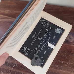 May include: An open book displaying a black Ouija board and a planchette. The board features white lettering and symbols, including the alphabet, numbers, and the words "yes," "no," and "goodbye." The planchette is dark grey.