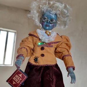 May include: A handmade doll with a blue face, round spectacles, and white hair. The doll is dressed in an orange jacket and a maroon skirt, holding a red book titled "Holy Bible". A small spider is attached to the jacket.