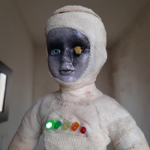 May include: A distinctive doll with a grey face, one blue eye, and one yellow eye. The doll is wrapped in white bandages and has a series of colourful lights on its chest. The doll's face has a distressed appearance.