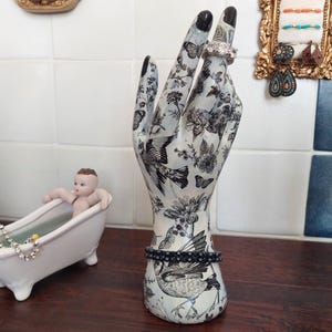 May include: A decorative hand-shaped jewellery holder with a black and white floral and bird pattern. A silver ring is on the index finger, and a black beaded bracelet is at the wrist. The fingernails are painted black.